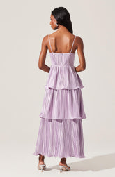 A woman is wearing a light purple, tiered maxi dress with vertical pinstripes. The dress features thin spaghetti straps and has four distinct layers, each with a ruffled edge. She is also wearing white high-heeled sandals and has short, natural hair.